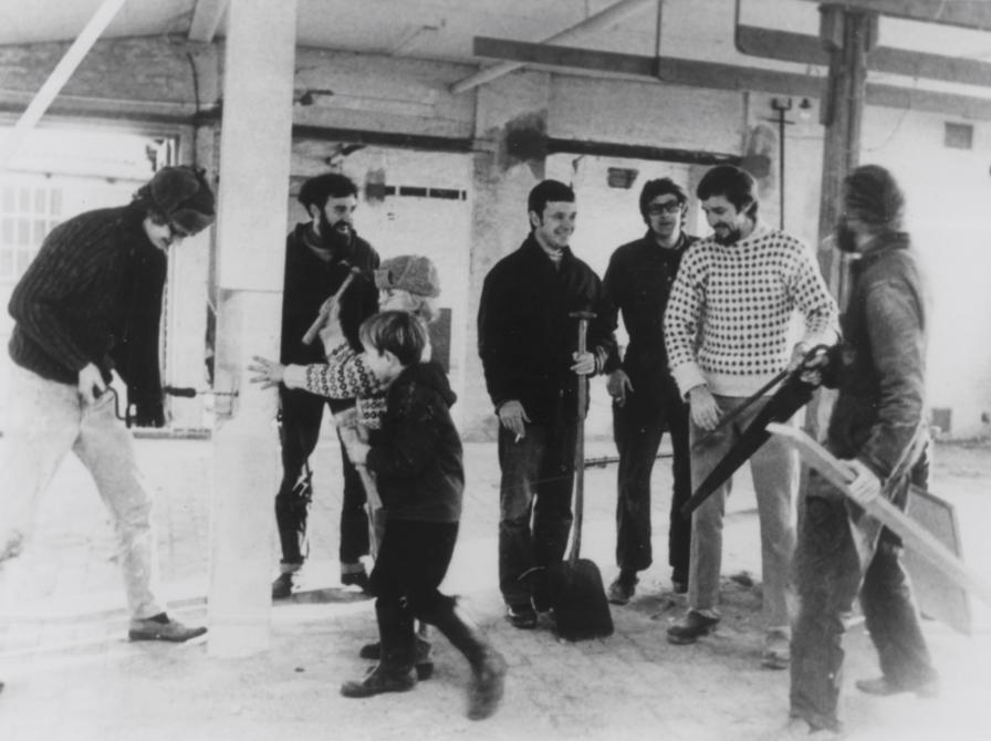 Sort-hvid foto af seks voksne og to børn i tom lagerhal. Alle smilende.