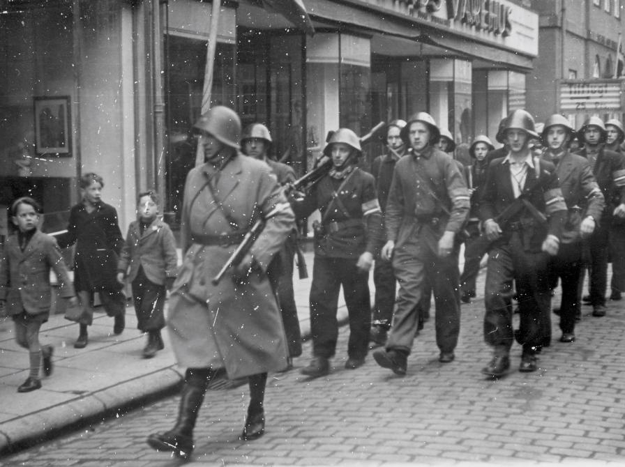 Sort-hvid foto af en gruppe modstandsfolk marcherende i række med børn på fortovet, der ser til.