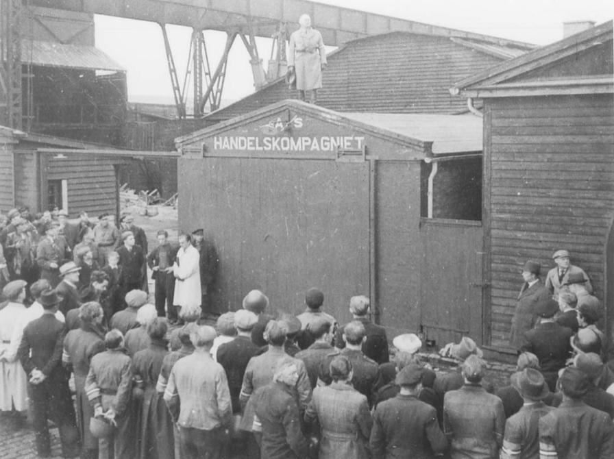 Sort-hvid foto af officer på taget af en havnebygning med tilhørere foran.